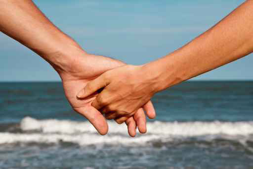 Das Bild zeigt zwei Personen die Händchen halten. Der Bildauschnitt reicht bis jeweils zur Mitte des Unterarmes. Im Hintergrund ist verschwommen eine Aussicht auf eine Welle am Ufer zu erkennen.